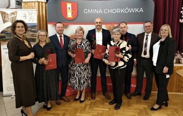 Grupa dziewięciu os&oacute;b zgromadzonych na ściance promocyjnej Gminy Chork&oacute;wka. Pięć os&oacute;b w środku trzyma czerwone foldery/dyplomy oraz kwiaty. Po bokach stoją przedstawiciele władz lokalnych oraz organizatorzy wydarzenia.