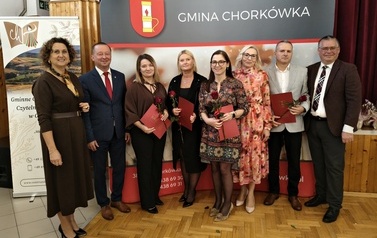 Osiem os&oacute;b pozuje na tle ścianki wystawienniczej Gminy Chork&oacute;wka. Wyr&oacute;żnione osoby trzymają czerwone teczki z nagrodami i czerwoną r&oacute;żę. W grupie znajdują się zar&oacute;wno kobiety, jak i mężczyzna odbierający wyr&oacute;żnienie.