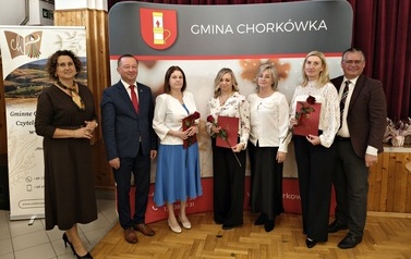 Pamiątkowa fotografia grupy siedmiu os&oacute;b na tle ścianki z herbem i napisem &bdquo;Gmina Chork&oacute;wka&rdquo;. Cztery kobiety stojące w środku trzymają w rękach czerwone dyplomy oraz r&oacute;że. Z obu stron towarzyszą im reprezentanci samorządu w formalnych strojach.