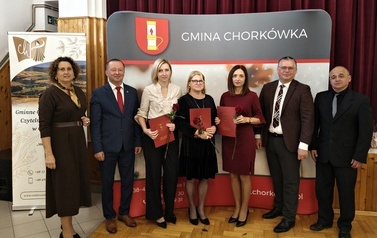 Pamiątkowe zdjęcie siedmiorga uczestnik&oacute;w uroczystości na tle ścianki Gminy Chork&oacute;wka. Trzy kobiety stojące w centrum trzymają czerwone dyplomy w teczkach oraz czerwoną r&oacute;żę. Po lewej i prawej stronie stoją przedstawiciele gminy w garniturach oraz prowadząca uroczystość.