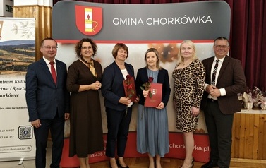 Grupa sześciu os&oacute;b pozuje do pamiątkowego zdjęcia na tle ścianki promocyjnej &bdquo;Gmina Chork&oacute;wka&rdquo;. W środku dwie kobiety trzymają czerwone pamiątkowe teczki oraz pojedyncze czerwone r&oacute;że. Po bokach stoją przedstawiciele władz gminy i organizatorzy w strojach wizytowych.