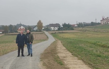 Dwaj mężczyźni stoją na środku wiejskiej drogi utwardzonej kruszywem, uśmiechając się do aparatu. Mężczyzna po lewej ma na sobie czarny płaszcz, a mężczyzna po prawej &ndash; oliwkową kurtkę bomberkę i jasne dżinsy. Droga prowadzi w głąb kadru, w stronę oddalonej wioski z kilkoma domami i widocznym kościołem na wzg&oacute;rzu po prawej stronie. Otoczenie stanowią pola uprawne i łąki pod pochmurnym, szarym niebem. Jest jesień, drzewa są częściowo bez liści.