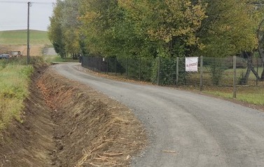 Wąska, kręta droga szutrowa prowadzi w d&oacute;ł zbocza, zakręcając w lewo. Po lewej stronie rozciąga się zielona łąka, a dalej widać zabudowania wiejskie i gospodarstwo. Po prawej stronie drogi rośnie gęsty pas wysokich, smukłych drzew (prawdopodobnie topoli) bez liści. Na pierwszym planie po prawej suche trawy. Krajobraz jest pag&oacute;rkowaty, pod ponurym, pochmurnym niebem.