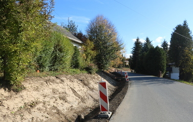 Prace budowlane przy drodze powiatowej polegające na budowie nowego chodnika oraz profilowaniu i umacnianiu row&oacute;w odwadniających. Na zdjęciach widoczne są odcinki drogi z ustawionymi biało-czerwonymi pachołkami ostrzegawczymi, wykopana i wyr&oacute;wnana ziemia pod krawężniki i podbudowę, a także fragmenty row&oacute;w wyłożone ażurowymi płytami betonowymi. W tle widoczne są domy jednorodzinne, zielone oraz jesienne drzewa i znaki drogowe.