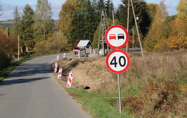 Prace budowlane przy drodze powiatowej polegające na budowie nowego chodnika oraz profilowaniu i umacnianiu row&oacute;w odwadniających. Na zdjęciach widoczne są odcinki drogi z ustawionymi biało-czerwonymi pachołkami ostrzegawczymi, wykopana i wyr&oacute;wnana ziemia pod krawężniki i podbudowę, a także fragmenty row&oacute;w wyłożone ażurowymi płytami betonowymi. W tle widoczne są domy jednorodzinne, zielone oraz jesienne drzewa i znaki drogowe.