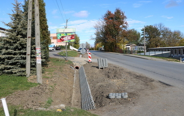 Prace budowlane przy drodze powiatowej polegające na budowie nowego chodnika oraz profilowaniu i umacnianiu row&oacute;w odwadniających. Na zdjęciach widoczne są odcinki drogi z ustawionymi biało-czerwonymi pachołkami ostrzegawczymi, wykopana i wyr&oacute;wnana ziemia pod krawężniki i podbudowę, a także fragmenty row&oacute;w wyłożone ażurowymi płytami betonowymi. W tle widoczne są domy jednorodzinne, zielone oraz jesienne drzewa i znaki drogowe.