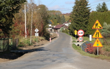Prace budowlane przy drodze powiatowej polegające na budowie nowego chodnika oraz profilowaniu i umacnianiu row&oacute;w odwadniających. Na zdjęciach widoczne są odcinki drogi z ustawionymi biało-czerwonymi pachołkami ostrzegawczymi, wykopana i wyr&oacute;wnana ziemia pod krawężniki i podbudowę, a także fragmenty row&oacute;w wyłożone ażurowymi płytami betonowymi. W tle widoczne są domy jednorodzinne, zielone oraz jesienne drzewa i znaki drogowe.