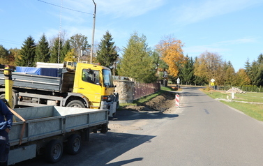 Prace budowlane przy drodze powiatowej polegające na budowie nowego chodnika oraz profilowaniu i umacnianiu row&oacute;w odwadniających. Na zdjęciach widoczne są odcinki drogi z ustawionymi biało-czerwonymi pachołkami ostrzegawczymi, wykopana i wyr&oacute;wnana ziemia pod krawężniki i podbudowę, a także fragmenty row&oacute;w wyłożone ażurowymi płytami betonowymi. W tle widoczne są domy jednorodzinne, zielone oraz jesienne drzewa i znaki drogowe.