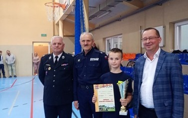 Zdjęcia przedstawiają uczestnik&oacute;w w strojach sportowych i strażackich podczas rywalizacji na hali sportowej, m.in. dynamiczne konkurencje z wężami strażackimi, uroczystą zbi&oacute;rkę wszystkich drużyn, a także momenty wręczania puchar&oacute;w, dyplom&oacute;w oraz nagr&oacute;d indywidualnych przez zaproszonych gości i przedstawicieli straży pożarnej.