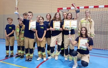Zdjęcia przedstawiają uczestnik&oacute;w w strojach sportowych i strażackich podczas rywalizacji na hali sportowej, m.in. dynamiczne konkurencje z wężami strażackimi, uroczystą zbi&oacute;rkę wszystkich drużyn, a także momenty wręczania puchar&oacute;w, dyplom&oacute;w oraz nagr&oacute;d indywidualnych przez zaproszonych gości i przedstawicieli straży pożarnej.