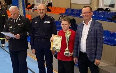 Zdjęcia przedstawiają uczestnik&oacute;w w strojach sportowych i strażackich podczas rywalizacji na hali sportowej, m.in. dynamiczne konkurencje z wężami strażackimi, uroczystą zbi&oacute;rkę wszystkich drużyn, a także momenty wręczania puchar&oacute;w, dyplom&oacute;w oraz nagr&oacute;d indywidualnych przez zaproszonych gości i przedstawicieli straży pożarnej.