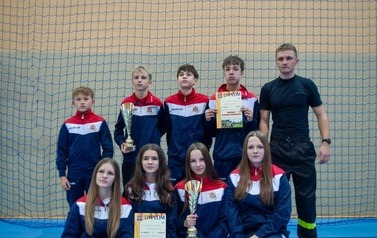 Zdjęcia przedstawiają uczestnik&oacute;w w strojach sportowych i strażackich podczas rywalizacji na hali sportowej, m.in. dynamiczne konkurencje z wężami strażackimi, uroczystą zbi&oacute;rkę wszystkich drużyn, a także momenty wręczania puchar&oacute;w, dyplom&oacute;w oraz nagr&oacute;d indywidualnych przez zaproszonych gości i przedstawicieli straży pożarnej.