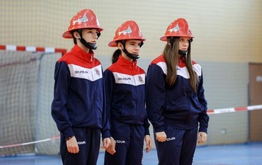Zdjęcia przedstawiają uczestnik&oacute;w w strojach sportowych i strażackich podczas rywalizacji na hali sportowej, m.in. dynamiczne konkurencje z wężami strażackimi, uroczystą zbi&oacute;rkę wszystkich drużyn, a także momenty wręczania puchar&oacute;w, dyplom&oacute;w oraz nagr&oacute;d indywidualnych przez zaproszonych gości i przedstawicieli straży pożarnej.