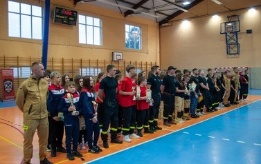 Zdjęcia przedstawiają uczestnik&oacute;w w strojach sportowych i strażackich podczas rywalizacji na hali sportowej, m.in. dynamiczne konkurencje z wężami strażackimi, uroczystą zbi&oacute;rkę wszystkich drużyn, a także momenty wręczania puchar&oacute;w, dyplom&oacute;w oraz nagr&oacute;d indywidualnych przez zaproszonych gości i przedstawicieli straży pożarnej.