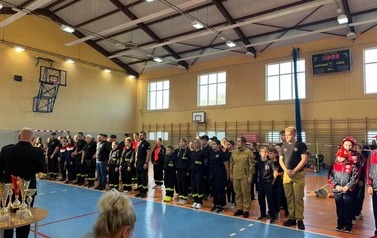 Zdjęcia przedstawiają uczestnik&oacute;w w strojach sportowych i strażackich podczas rywalizacji na hali sportowej, m.in. dynamiczne konkurencje z wężami strażackimi, uroczystą zbi&oacute;rkę wszystkich drużyn, a także momenty wręczania puchar&oacute;w, dyplom&oacute;w oraz nagr&oacute;d indywidualnych przez zaproszonych gości i przedstawicieli straży pożarnej.