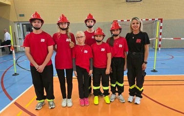 Zdjęcia przedstawiają uczestnik&oacute;w w strojach sportowych i strażackich podczas rywalizacji na hali sportowej, m.in. dynamiczne konkurencje z wężami strażackimi, uroczystą zbi&oacute;rkę wszystkich drużyn, a także momenty wręczania puchar&oacute;w, dyplom&oacute;w oraz nagr&oacute;d indywidualnych przez zaproszonych gości i przedstawicieli straży pożarnej.