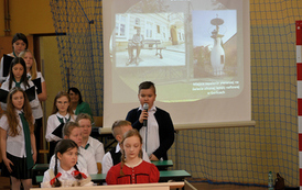 Uczniowie, w strojach dawnych siedzą w ławkach i odgrywają scenki rodzajowe - szkolne życie. 