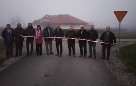 Otwarcie drogi. Grupa ludzi stoi w poprzek drogi przed szarfą, kt&oacute;ra ma być przecięta jako symbol oddania nowej drogi do użytku.   