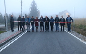 Otwarcie drogi. Grupa ludzi stoi w poprzek drogi przed szarfą, kt&oacute;ra ma być przecięta jako symbol oddania nowej drogi do użytku.