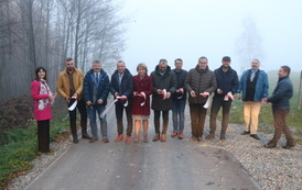 Otwarcie drogi. Grupa ludzi stoi w poprzek drogi przed szarfą, kt&oacute;ra ma być przecięta jako symbol oddania nowej drogi do użytku.