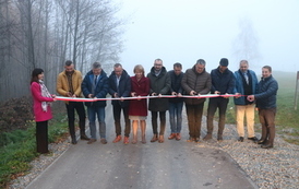 Otwarcie drogi. Grupa ludzi stoi w poprzek drogi przed szarfą, kt&oacute;ra ma być przecięta jako symbol oddania nowej drogi do użytku.