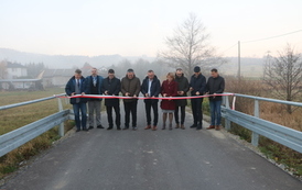 Otwarcie drogi. Grupa ludzi stoi w poprzek drogi przed szarfą, kt&oacute;ra ma być przecięta jako symbol oddania nowej drogi do użytku.