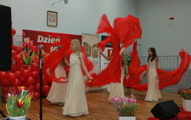 Dziewczyny w beżowych sukienkach trzymające w dłoniach czerwone wachlarze oraz czerwone zwiewne pasma materiał&oacute;w. Tanczą układ z wachlarzami. 