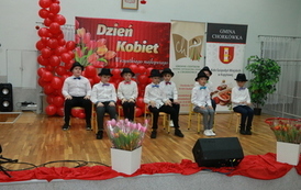 Chłopcy w białych koszulach i kapeluszach, tańczą przy krzesłach. 