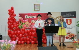 Dw&oacute;ch wokalist&oacute;w w białych koszulach oraz wokalistka w ż&oacute;łtej sukience. Śpiewają. 