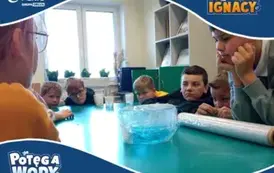 Grupa chłopc&oacute;w z zainteresowaniem obserwuje eksperyment z wodą. Na środku stołu znajduje się miska owinięta folią spożywczą z niebieską cieczą w środku. Uczniowie są skupieni i zaciekawieni. Na grafice umieszczono loga &bdquo;Fundacja PGNiG Grupa Orlen&rdquo;, &bdquo;Być j