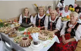 Grupa kobiet w tradycyjnych ludowych strojach siedzi przy bogato zastawionym stole, pełnym kolorowych dań wielkanocnych, ciast i dekoracji. Kobiety mają na sobie białe bluzki z koronkowymi kołnierzykami, ciemne gorsety i czerwone sp&oacute;dnice, a ich szyje zdo