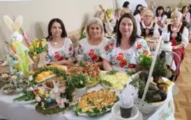 Grupa kobiet w tradycyjnych ludowych strojach siedzi przy bogato zastawionym stole, pełnym kolorowych dań wielkanocnych, ciast i dekoracji. Kobiety mają na sobie białe bluzki z koronkowymi kołnierzykami, ciemne gorsety i czerwone sp&oacute;dnice, a ich szyje zdo