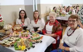 Grupa kobiet w tradycyjnych ludowych strojach siedzi przy bogato zastawionym stole, pełnym kolorowych dań wielkanocnych, ciast i dekoracji. Kobiety mają na sobie białe bluzki z koronkowymi kołnierzykami, ciemne gorsety i czerwone sp&oacute;dnice, a ich szyje zdo