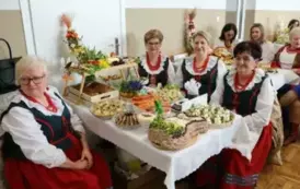 Grupa kobiet w tradycyjnych ludowych strojach siedzi przy bogato zastawionym stole, pełnym kolorowych dań wielkanocnych, ciast i dekoracji. Kobiety mają na sobie białe bluzki z koronkowymi kołnierzykami, ciemne gorsety i czerwone sp&oacute;dnice, a ich szyje zdo