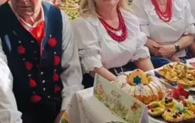 Grupa kobiet w tradycyjnych ludowych strojach siedzi przy bogato zastawionym stole, pełnym kolorowych dań wielkanocnych, ciast i dekoracji. Kobiety mają na sobie białe bluzki z koronkowymi kołnierzykami, ciemne gorsety i czerwone sp&oacute;dnice, a ich szyje zdo