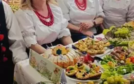 Grupa kobiet w tradycyjnych ludowych strojach siedzi przy bogato zastawionym stole, pełnym kolorowych dań wielkanocnych, ciast i dekoracji. Kobiety mają na sobie białe bluzki z koronkowymi kołnierzykami, ciemne gorsety i czerwone sp&oacute;dnice, a ich szyje zdo