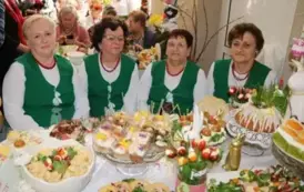Grupa kobiet w tradycyjnych ludowych strojach siedzi przy bogato zastawionym stole, pełnym kolorowych dań wielkanocnych, ciast i dekoracji. Kobiety mają na sobie białe bluzki z koronkowymi kołnierzykami, ciemne gorsety i czerwone sp&oacute;dnice, a ich szyje zdo