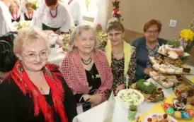 Grupa kobiet w tradycyjnych ludowych strojach siedzi przy bogato zastawionym stole, pełnym kolorowych dań wielkanocnych, ciast i dekoracji. Kobiety mają na sobie białe bluzki z koronkowymi kołnierzykami, ciemne gorsety i czerwone sp&oacute;dnice, a ich szyje zdo