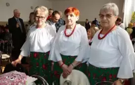 Grupa kobiet w tradycyjnych ludowych strojach siedzi przy bogato zastawionym stole, pełnym kolorowych dań wielkanocnych, ciast i dekoracji. Kobiety mają na sobie białe bluzki z koronkowymi kołnierzykami, ciemne gorsety i czerwone sp&oacute;dnice, a ich szyje zdo