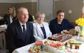 Grupa kobiet w tradycyjnych ludowych strojach siedzi przy bogato zastawionym stole, pełnym kolorowych dań wielkanocnych, ciast i dekoracji. Kobiety mają na sobie białe bluzki z koronkowymi kołnierzykami, ciemne gorsety i czerwone sp&oacute;dnice, a ich szyje zdo
