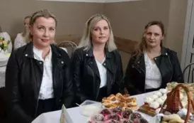 Grupa kobiet w tradycyjnych ludowych strojach siedzi przy bogato zastawionym stole, pełnym kolorowych dań wielkanocnych, ciast i dekoracji. Kobiety mają na sobie białe bluzki z koronkowymi kołnierzykami, ciemne gorsety i czerwone sp&oacute;dnice, a ich szyje zdo