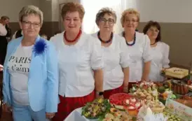 Grupa kobiet w tradycyjnych ludowych strojach siedzi przy bogato zastawionym stole, pełnym kolorowych dań wielkanocnych, ciast i dekoracji. Kobiety mają na sobie białe bluzki z koronkowymi kołnierzykami, ciemne gorsety i czerwone sp&oacute;dnice, a ich szyje zdo