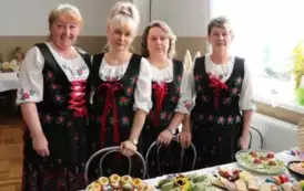 Grupa kobiet w tradycyjnych ludowych strojach siedzi przy bogato zastawionym stole, pełnym kolorowych dań wielkanocnych, ciast i dekoracji. Kobiety mają na sobie białe bluzki z koronkowymi kołnierzykami, ciemne gorsety i czerwone sp&oacute;dnice, a ich szyje zdo