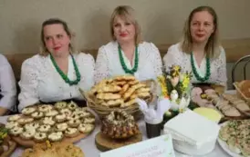 Grupa kobiet w tradycyjnych ludowych strojach siedzi przy bogato zastawionym stole, pełnym kolorowych dań wielkanocnych, ciast i dekoracji. Kobiety mają na sobie białe bluzki z koronkowymi kołnierzykami, ciemne gorsety i czerwone sp&oacute;dnice, a ich szyje zdo