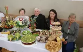 Grupa kobiet w tradycyjnych ludowych strojach siedzi przy bogato zastawionym stole, pełnym kolorowych dań wielkanocnych, ciast i dekoracji. Kobiety mają na sobie białe bluzki z koronkowymi kołnierzykami, ciemne gorsety i czerwone sp&oacute;dnice, a ich szyje zdo