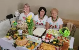Grupa kobiet w tradycyjnych ludowych strojach siedzi przy bogato zastawionym stole, pełnym kolorowych dań wielkanocnych, ciast i dekoracji. Kobiety mają na sobie białe bluzki z koronkowymi kołnierzykami, ciemne gorsety i czerwone sp&oacute;dnice, a ich szyje zdo