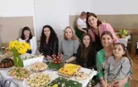 Grupa kobiet w tradycyjnych ludowych strojach siedzi przy bogato zastawionym stole, pełnym kolorowych dań wielkanocnych, ciast i dekoracji. Kobiety mają na sobie białe bluzki z koronkowymi kołnierzykami, ciemne gorsety i czerwone sp&oacute;dnice, a ich szyje zdo