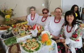 Grupa kobiet w tradycyjnych ludowych strojach siedzi przy bogato zastawionym stole, pełnym kolorowych dań wielkanocnych, ciast i dekoracji. Kobiety mają na sobie białe bluzki z koronkowymi kołnierzykami, ciemne gorsety i czerwone sp&oacute;dnice, a ich szyje zdo