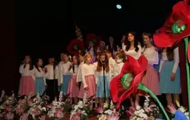 Grupa dorosłych os&oacute;b stoi razem na scenie, pozując do pamiątkowego zdjęcia. Większość z nich trzyma w rękach r&oacute;że. W tle czarna kurtyna i niebieskie światła sceniczne. Na pierwszym planie widoczne są kompozycje kwiatowe. Atmosfera wskazuje na oficjalne wy