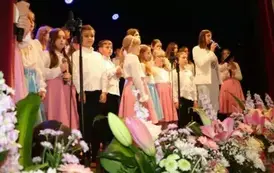 Grupa dorosłych os&oacute;b stoi razem na scenie, pozując do pamiątkowego zdjęcia. Większość z nich trzyma w rękach r&oacute;że. W tle czarna kurtyna i niebieskie światła sceniczne. Na pierwszym planie widoczne są kompozycje kwiatowe. Atmosfera wskazuje na oficjalne wy
